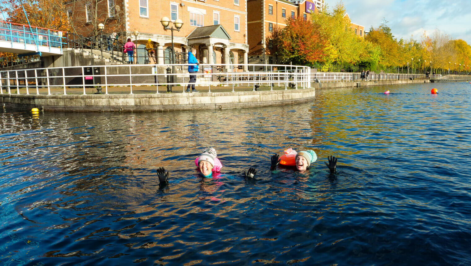 The image shows two swimmers waving to the camera on one of the cold water swimming sessions. The sun is shining.